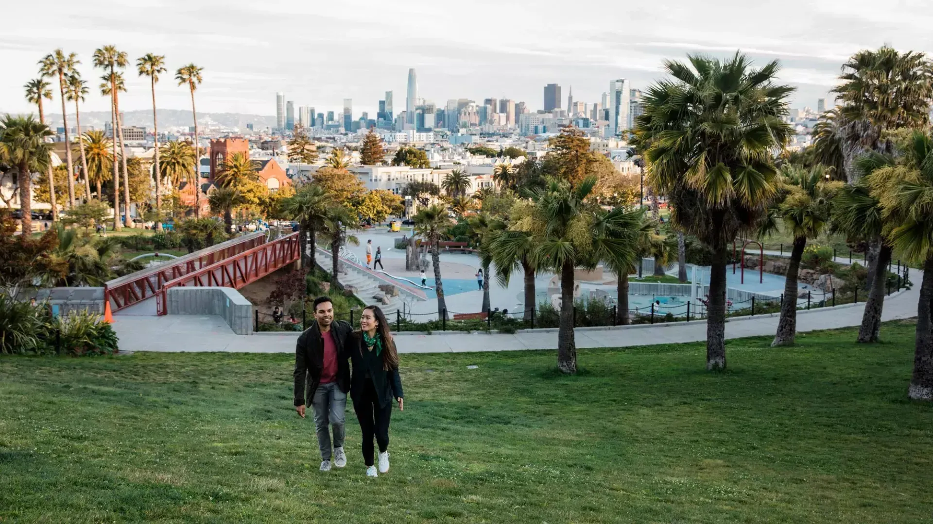 A couple walks toward the camera with Dolores Park and the San Francisco Skyline behind them.